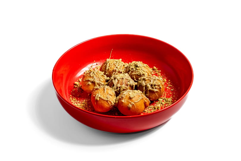 Six golden-brown donut holes drizzled with pistachio sauce and sprinkled with crushed pistachios in a red bowl.