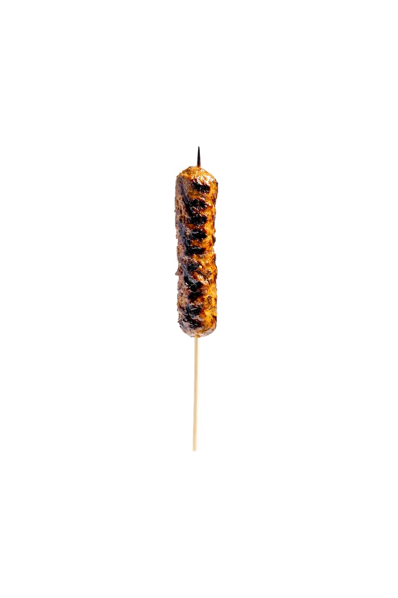 A single grilled meat skewer, golden brown and charred, stands upright on a white background.