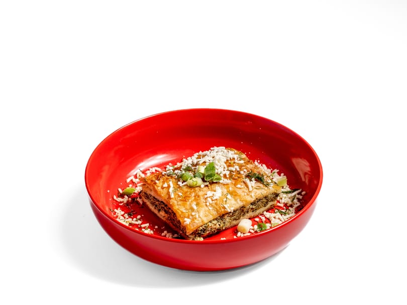 A golden-brown savory pastry, possibly spanakopita, in a red bowl, garnished with herbs and feta cheese.