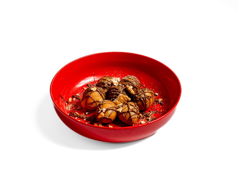 Six golden-brown donut holes drizzled with chocolate and sprinkled with nuts in a red bowl.